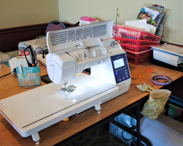 A white and blue NQ900 sewing machine is shown set up on a brown table with an assortment of sewing tools and notions on its right side. Brother NQ900 sewing machine