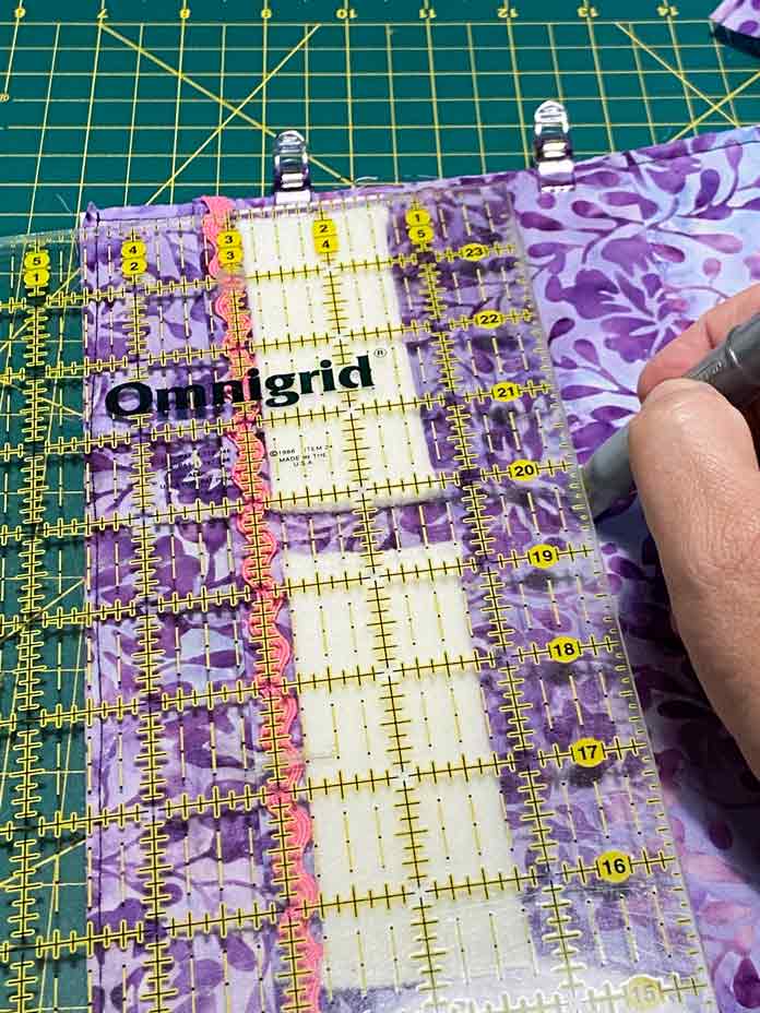 A hand holding a silver Chaco liner marker is shown drawing a line along the edge of a yellow gridded quilting ruler. Purple fabric, white wool rectangles and pink rick rack can be seen through the ruler. Purple Clever Clips hold the layers of fabric together. Brother NQ900 sewing machine, UNIQUE quilting Clever Clips, UNIQUE Medium Rick Rack