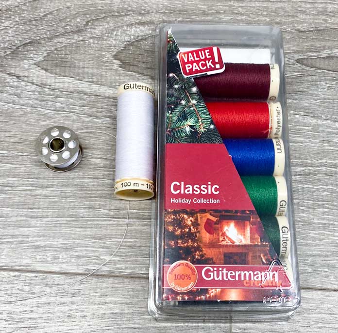 A package of Gütermann Classic Holiday Collection thread is next to a spool of white thread and a metal bobbin with the same white thread.