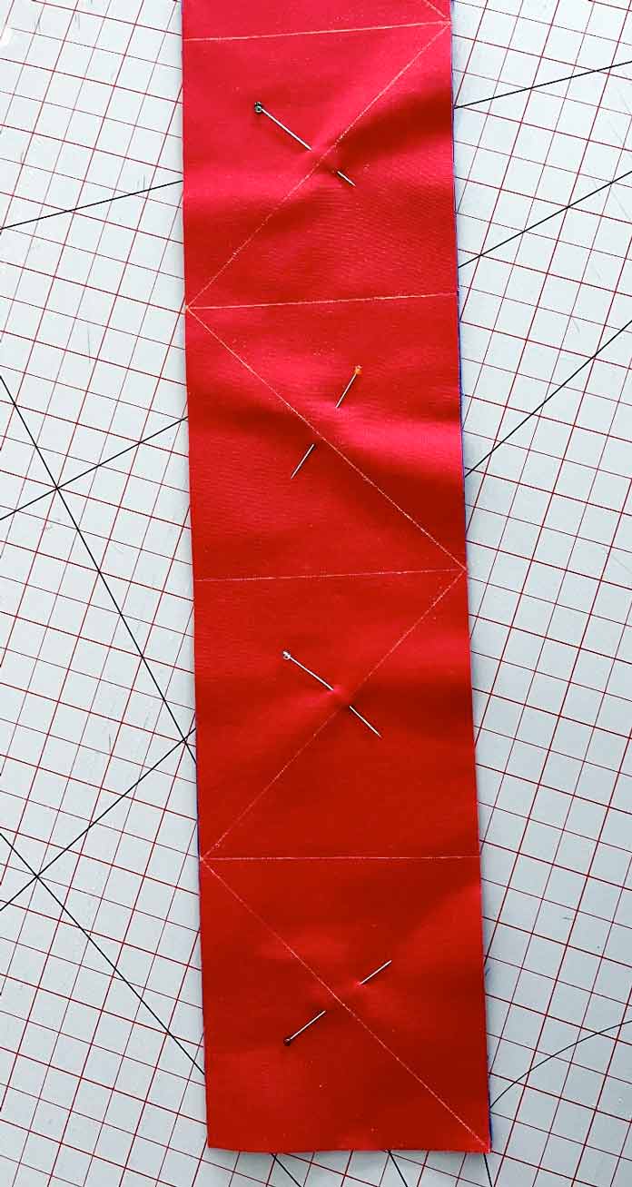 A strip of red fabric is pinned and marked with horizontal and diagonal lines. Clover Chaco Liner, Clover Quilting Pins, Clover Rotary Cutter