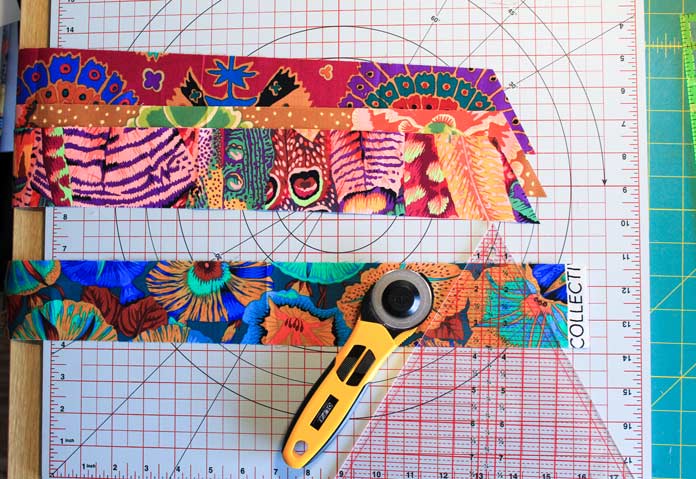 2½” strips of Kaffe Fasset fabrics with one edge trimmed at a 60-degree angle lay on a cutting mat next to a triangle ruler and rotary cutter; Komfort Kut 360° Rotating Cutting Mat - 18" x 18", Sew Easy Triangle Ruler 60°, OLFA Quick Change Rotary Cutter