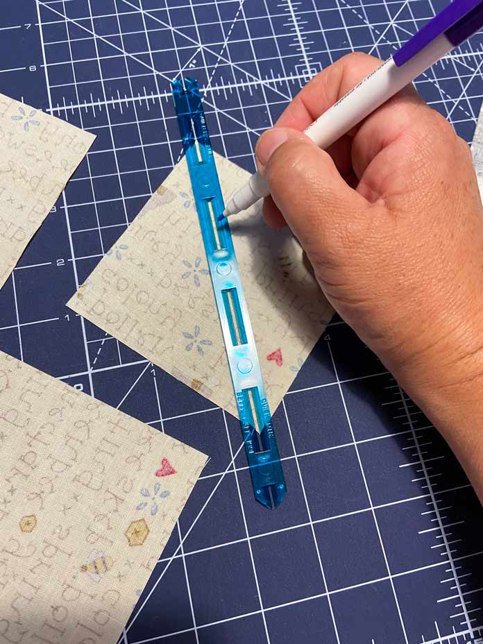 A hand is shown using a blue ruler and a white fabric pen to draw a diagonal line on the back of one square of yellow fabric. Portions of two other squares can be seen on the left side of the photo and all of the fabric squares are on a blue cutting mat; OLFA Double Sided Cutting Mat (navy blue)