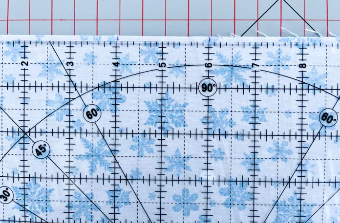Marks placed at an equidistance from each edge of a white fabric with a blue snowflake print; a quilting ruler is on top of the fabric that lies on a gray cutting mat with red grid lines; OLFA 12½″ Square Frosted Acrylic Ruler, OLFA 6″ x 12″ Frosted Acrylic Ruler, Heirloom 24″ x 36″ Double Sided Cutting Mat