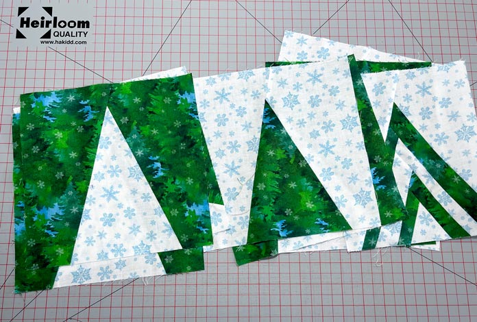 Nine tree quilt blocks in white blue and green fabrics are trimmed to a 9” square and lay on top of each other on a gray cutting mat with red grid lines