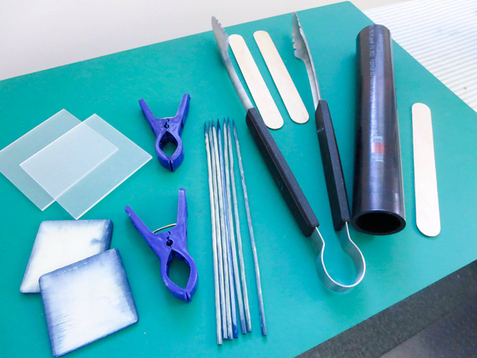 Extra tools for dyeing set against a green background include 2 plastic squares, 2 small wooden squares, 2 clothes pins, 7 wooden skewers, a pair of tongs, black PVC pipe, 4 craft sticks 