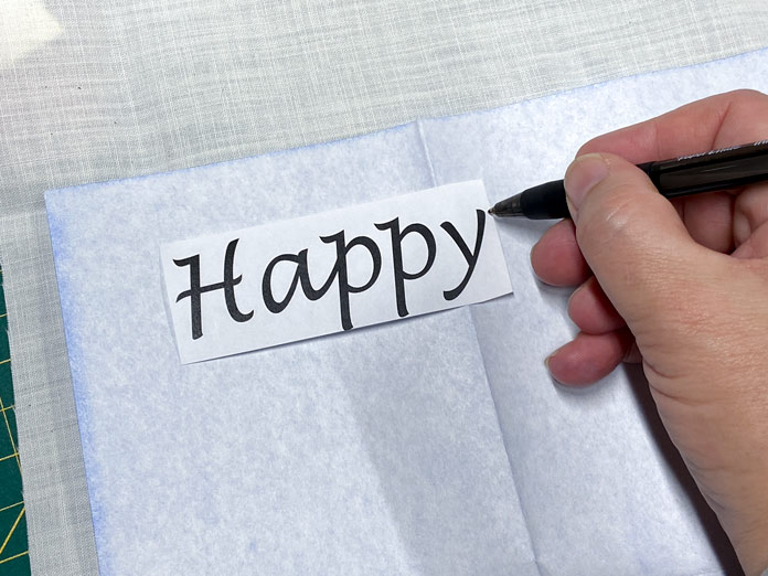 A hand traces the word Happy onto a cream-colored piece of fabric using a black ballpoint pen and a piece of blue transfer paper.