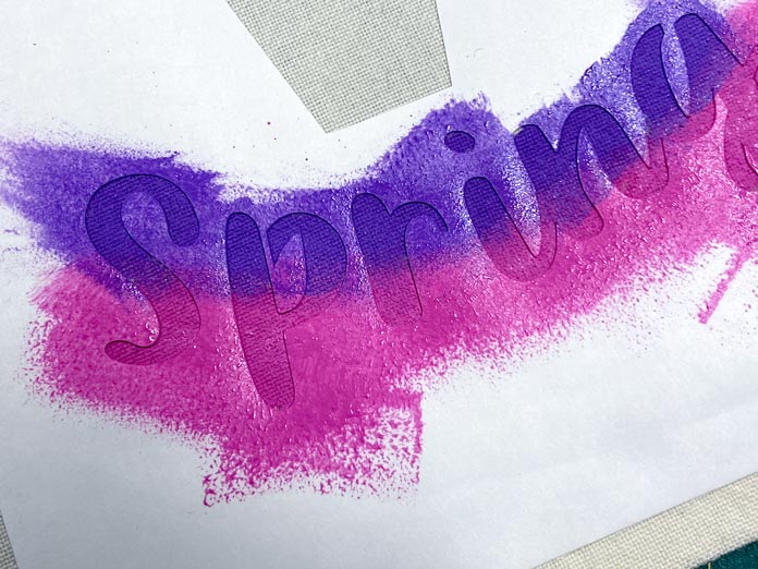 Pink and purple paint is shown on top of a white stencil of the word “spring” that has been ironed onto a beige tea towel; Sew Easy Freezer Paper for Quilting and Applique, Mont Marte Signature Fabric Paints
