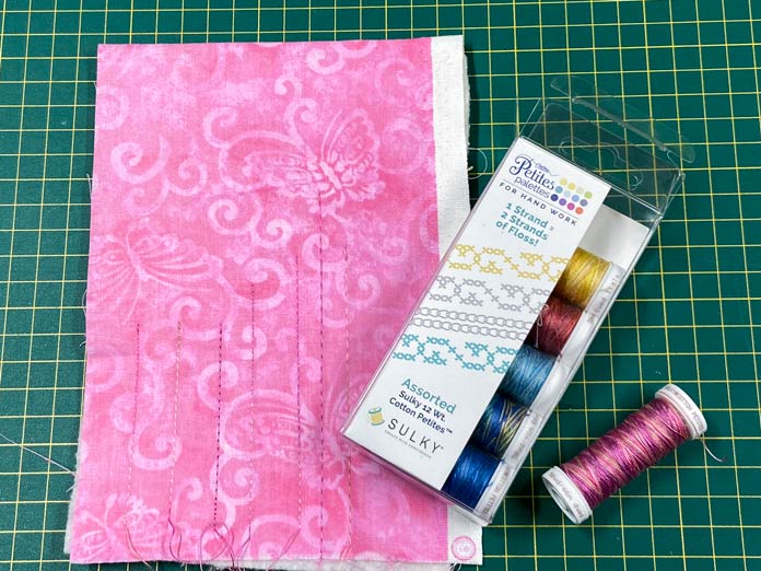 a small piece of pink fabric with stitched lines in pink thread and a box of Sulky Cotton Petites 6 Spool Thread Set on a green cutting mat