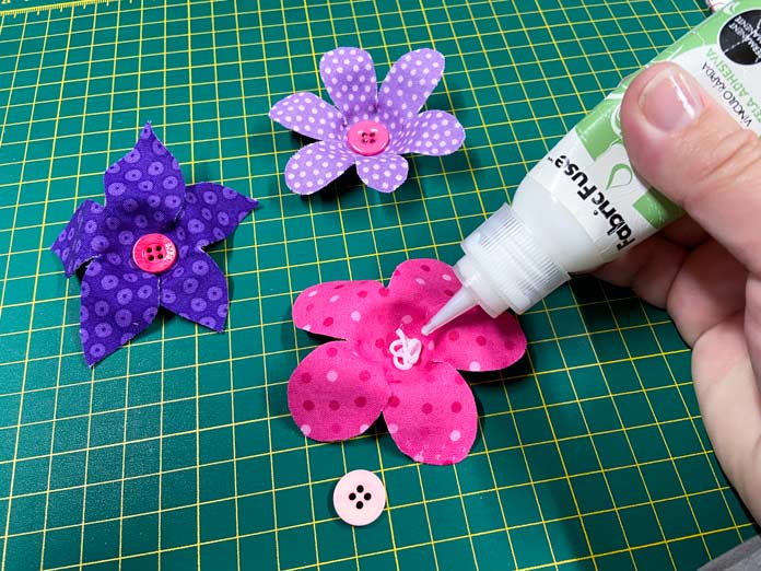 A hand is shown squeezing adhesive from a small white and green bottle of Fabric Fuse by HeatnBond into the middle of a pink 3D fabric flower. Two other flowers are also shown sitting on the green cutting mat in the background; Crafting Essentials Bottle of Buttons - Pink Tones - 75g (2.6oz), Fabric Fuse by HeatnBond Quick Bond Fabric Adhesive