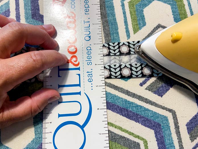 A hand is shown holding a ruler on top of a strip of fabric. A yellow iron is pressing the fabric strip.