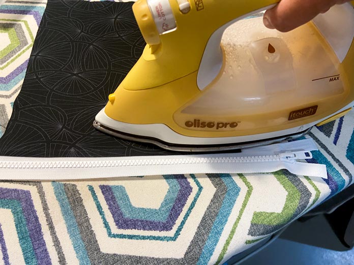 A yellow iron is shown pressing a black fabric with a white zipper sewn to it on top of a multicolored ironing board.