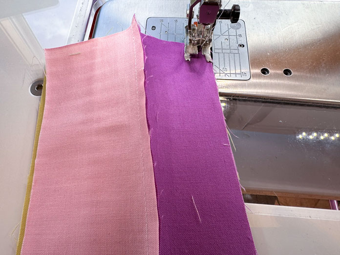 A pink and purple fabric strip are sewn together and under a sewing machine foot and needle ready to be sewn along the right edge. Hemline Gold Multi-Use Craft Bag (28.5cm x 36.5cm x 17.8cm), Hemline Gold Sewing Kit, Hemline Gold Quilters 4-in-1 Multi-Mat (30cm x 24cm), Hemline Gold Quilters Clips (Pack of 30), Hemlione Gold Retractable Tape Measure - 150cm/60, Oliso M3Pro Project Iron – Coral, OLFA 45mm RTY-2/DX/MAG Ergonomic Rotary Cutter – Magenta, OLFA RM-MG - 24″ x 36″ Double Sided Rotary Mat, UNIQUE Sewing Tools Holder, OLFA QR-6x12 - 6″ x 12″ Frosted Acrylic Ruler, OLFA QR-6x24 - 6″ x 24″ Frosted Acrylic Ruler, Fairfield Quilter's 80/20 Quilt Batting - 229 x 274cm (90″ x 108″)