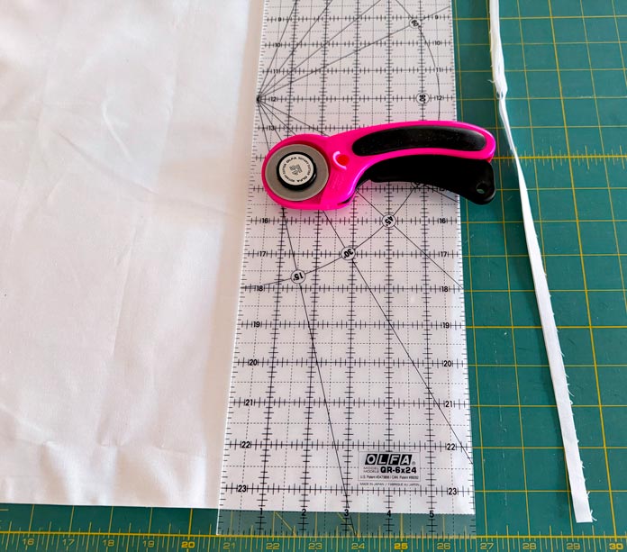 A pink rotary cutter rests on top of an OLFA ruler, placed on the folded edge of a white fabric on a green cutting mat. The white fabric has been trimmed to straighten one edge. Hemline Gold Multi-Use Craft Bag (28.5cm x 36.5cm x 17.8cm), Hemline Gold Sewing Kit, Hemline Gold Quilters 4-in-1 Multi-Mat (30cm x 24cm), Hemline Gold Quilters Clips (Pack of 30), Hemline Gold Retractable Tape Measure - 150cm/60, oliso M3Pro Project Iron – Coral, OLFA 45mm RTY-2/DX/MAG Ergonomic Rotary Cutter – Magenta, OLFA RM-MG - 24″ x 36″ Double Sided Rotary Mat, UNIQUE Sewing Tools Holder, OLFA QR-6x12 - 6″ x 12″ Frosted Acrylic Ruler, OLFA QR-6x24 - 6″ x 24″ Frosted Acrylic Ruler, Fairfield Quilter's 80/20 Quilt Batting - 229 x 274cm (90″ x 108″)