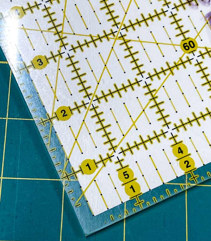A yellow gridded Omnigrid Ruler is shown on a green cutting mat with a piece of white paper-backed Clear Grip underneath it. UNIQUE Clear Grip, UNIQUE quilting Ruler/Template Grips, Omnigrid Ruler, Omnigrid Triangle Ruler, Fabric Creations Fabric Palette Pre-Cut Fabrics, Fairfield Crafter’s Choice Pillow Form