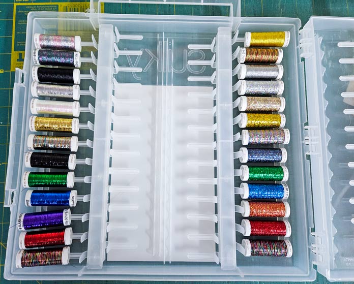 A variety of colored metallic threads in a clear plastic box with a green cutting mat in the background. Husqvarna VIKING DESIGNER EPIC 3 sewing and embroidery machine, Husqvarna VIKING 6in/15.2cm Applique Scissor, Husqvarna VIKING Open Toe Presser Foot for IDF System, SULKY Slimline Metallic Dream Assortment