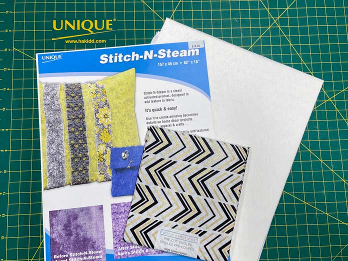 A white sheet of Stitch-N-Steam is shown under the blue, yellow and white package. A fat quarter of white fabric with a black and gold zig-zag pattern sits on top with a green cutting mat in the background. Oliso M2Pro Mini Project Iron, FAIRFIELD Low-Loft Quilt Batting - Crib - 114 x 152cm (45″ x 60″), UNIQUE QUILTING Clever Clips, OLFA CHN-1 - Chenille / Textile Cutter, UNIQUE CREATIV Trim Pack - Black & White, FABRIC CREATIONS Burlap Fabric - Oyster - 45 x 61cm (18″ x 24″), FABRIC CREATIONS Faux Fur Fabric - Zebra - 45 x 76cm (18″ x 30″), UNIQUE SEWING Double Adjust Parachute Buckle - Plastic - 25mm (1″) White, CRAFTING ESSENTIALS Bottle of Buttons - Pink Tones - 75g (2.6oz), UNIQUE SEWING Invisible Magnetic Snaps - Sew-On - 12mm (1⁄2″) - 2 sets, UNIQUE QUILTING Stitch-N-Steam - 157 x 45cm (62″ x 18″), FABRIC CREATIONS Fabric Bundle (5pcs) - Metallic Gold - 45 x 53cm (18in x 21in), Oliso Pro TG1600 Pro Plus Smart Iron, UNIQUE QUILTING Clever Clips Large - 12pcs, sewing tutorial