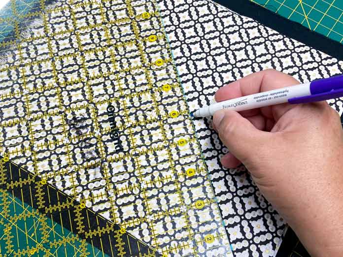 A hand draws a diagonal line on a black and white fabric using a yellow-gridded ruler and a purple-tipped fabric marker. Oliso M2Pro Mini Project Iron, FAIRFIELD Low-Loft Quilt Batting - Crib - 114 x 152cm (45″ x 60″), UNIQUE QUILTING Clever Clips, OLFA CHN-1 - Chenille / Textile Cutter, UNIQUE CREATIV Trim Pack - Black & White, FABRIC CREATIONS Burlap Fabric - Oyster - 45 x 61cm (18″ x 24″), FABRIC CREATIONS Faux Fur Fabric - Zebra - 45 x 76cm (18″ x 30″), UNIQUE SEWING Double Adjust Parachute Buckle - Plastic - 25mm (1″) White, CRAFTING ESSENTIALS Bottle of Buttons - Pink Tones - 75g (2.6oz), UNIQUE SEWING Invisible Magnetic Snaps - Sew-On - 12mm (1⁄2″) - 2 sets, UNIQUE QUILTING Stitch-N-Steam - 157 x 45cm (62″ x 18″), FABRIC CREATIONS Fabric Bundle (5pcs) - Metallic Gold - 45 x 53cm (18in x 21in), Oliso Pro TG1600 Pro Plus Smart Iron, UNIQUE QUILTING Clever Clips Large - 12pcs, UNIQUE SEWING 2-in-1 Dual-Tip Wash-out/Air Erasable Marking Pen - Fine, OMNIGRID Ruler - 121⁄2″ x 121⁄2″ (31.75 x 31.75cm), sewing tutorial