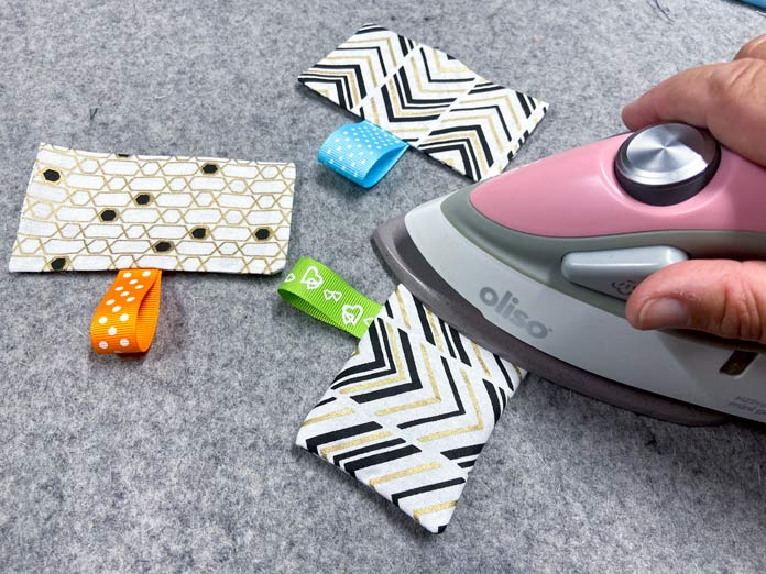 A hand holds a pink mini iron while pressing a white, gold, and black rectangle of fabric with a green ribbon loop attached. Two other rectangles of fabric with ribbons attached sit on top of the grey wool pressing mat in the background. OLFA CHN-1 - Chenille / Textile Cutter, UNIQUE QUILTING Stitch-N-Steam - 157 x 45cm (62″ x 18″), FABRIC CREATIONS Fabric Bundle (5pcs) - Metallic Gold - 45 x 53cm (18in x 21in), UNIQUE SEWING Self-Gripping Fastener Strip - 25cm (10″) - White, UNIQUE Webbing 25mm x 10m (1″ x 11yd) - White, an ELAN Trench Buckle - 45mm (13⁄4″) - Brown, UNIQUE CREATIV Trim Pack - Black & White, UNIQUE QUILTING Clever Clips Small - 12 pcs, CLOVER 7807 - Hold it Precision Stiletto, UNIQUE SEWING Sewing Tools Holder, UNIQUE QUILTING Wool Pressing Mat - 18″ x 24″ - Grey, OLISO M2Pro Mini Project Iron with Solemate, UNIQUE QUILTING Clever Clips Large - 12pcs, UNIQUE SEWING Invisible Magnetic Snaps - Sew-On - 12mm (1⁄2″) - 2 sets, COSTUMAKERS Activewear One Way Separating Zipper 23cm (9″) - White - 1760, UNIQUE SEWING Double Adjust Parachute Buckle - Plastic - 25mm (1″) - White, free tutorial