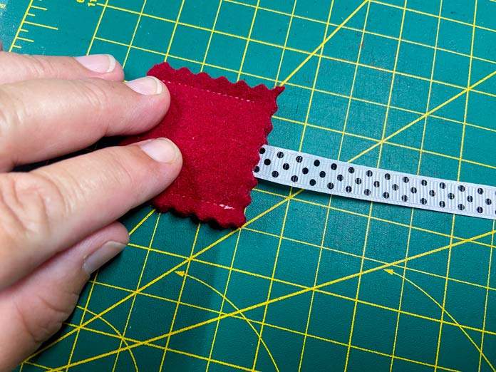 A hand holds a small pouch made of red wool felt. The end of a white ribbon with black polka dots is slipped into the open edge of the pouch. A green cutting mat is in the background. UNIQUE SEWING Self-Gripping Fastener Strip - 25cm (10″) - White, UNIQUE Webbing 25mm x 10m (1″ x 11yd) - White, ELAN Trench Buckle - 45mm (13⁄4″) - Brown, UNIQUE CREATIV Trim Pack - Black & White, UNIQUE SEWING Invisible Magnetic Snaps - Sew-On - 12mm (1⁄2″) - 2 sets,  SOFTKUT Pinking Shears (Scissors) - 81⁄2″ (21.6cm), COSTUMAKERS General Purpose Closed End Zipper 20cm (8″) - White - 1700, UNIQUE CREATIV Trim Pack - Red & Green, OLFA RTY-2/DX - Deluxe Ergonomic Handle Rotary Cutter 45mm, UNIQUE QUILTING Clever Clips Small - 12 pcs, UNIQUE SEWING Double Adjust Parachute Buckle - Plastic - 25mm (1″) - White, CRAFTING ESSENTIALS Bottle of Buttons - Pink Tones - 75g (2.6oz), UNIQUE Braided Elastic 3mm x 3.7m - White, free tutorial