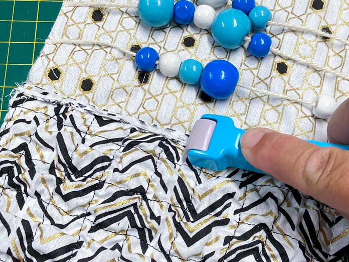 A hand uses a blue Roll & Press to open and flatten the seam between two black, white, and gold quilt blocks. Blue and white beads on white twine can be seen at the top of the picture. UNIQUE SEWING Invisible Magnetic Snaps - Sew-On - 12mm (1⁄2″) - 2 sets, COSTUMAKERS General Purpose Closed End Zipper 20cm (8″) - White - 1700, UNIQUE SEWING Double Adjust Parachute Buckle - Plastic - 25mm (1″) – White, UNIQUE QUILTING Clever Clips Small - 12 pcs, CLOVER 7812 Roll & Press, UNIQUE QUILTING Wool Pressing Mat - 18″ x 24″ - Grey, OLISO M2Pro Mini Project Iron with Solemate, CLOVER 240 - Fork Pins - .56mm, CLOVER 464/18 - Bias Tape Maker - 18mm (3⁄4″), Odif 505 Temporary Quilt Basting Adhesive Fabric Spray - 156g, FAIRFIELD Low-Loft® Quilt Batting - Crib - 114 x 152cm (45″ x 60″), free tutorial