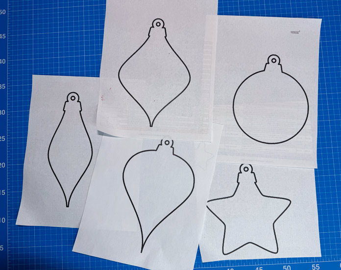 5 Christmas ornament shapes printed on paper lay on top of a blue cutting mat; Gütermann Dekor Metallic Thread 200m – Gold, Gütermann Dekor Metallic Thread 200m - Col. 41, Gütermann Dekor Metallic Thread 200m – Red, Gütermann Dekor Metallic Thread 200m - Christmas Green, Gütermann Dekor Metallic Thread 200m - Col. 1005, Gütermann Denim Thread 100m – Black, UNIQUE Baby Rick Rack 3mm x 3.7m – Scarlet, UNIQUE Medium Rick Rack 6mm x 2.3m - Emerald Green, UNIQUE Jumbo Rick Rack 10mm x 2.3m - Metallic Gold, SCHMETZ #1743 Metallic Needles Carded - 80/12 - 5 count, SCHMETZ #1752 Metallic Needles Carded - 90/14 - 5 count, LDH 8½“ Craft Scissors – Purple, HeatnBond Lite Iron-On Adhesive - 43cm x 30m (17″ x 32 ¾yds) bolt, HeatnBond Stitch n Sew Tear-Away Stabilizer - 50.8cm x 22.8m (20″ x 25yds), oliso M3Pro Project Iron – Coral, OLFA 45mm RTY-2/DX/MAG Ergonomic Rotary Cutter – Magenta, UNIQUE QUILTING Wool Pressing Mat - 18″ x 24″ - Grey 