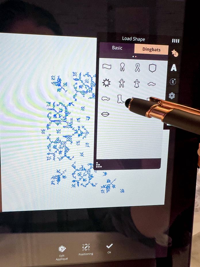 A purple model of the PFAFF creative icon 2 with Edit Applique mode with the Load Shape screen, category Dingbats; PFAFF creative icon 2, PFAFF Universal Needles Size 80/12, PFAFF Universal Needles Size 90/14, PFAFF Quilting Needles Size 90/14, PFAFF 6D Dynamic Spring Foot, PFAFF creative 120 Square Hoop (120 x 120 mm), Mettler 40wt polyester thread, PFAFF Aqua Magic Plus Stabilizer, PFAFF 0A Standard Presser Foot for IDT System 