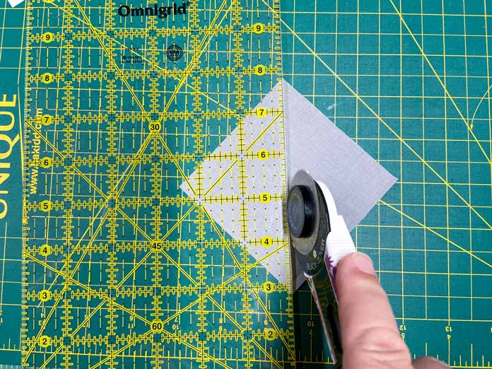 A hand uses a white rotary cutter and rotary cutting ruler to cut a gray square of fabric in half on top of a green cutting mat.