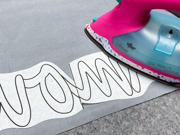 A pink iron is being used to iron a fusible applique design to the back of a gray piece of fabric. A mottled gray wool pressing mat is in the background.