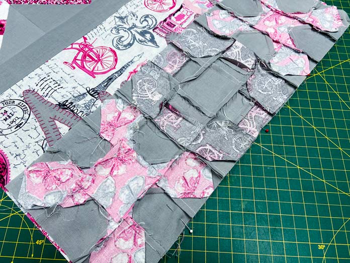 A pink, gray and white fabric rectangle sits on top of a green cutting mat. A row of pieced blocks made with gray and pink fabric has been pinned to the edge using red and white pins. 