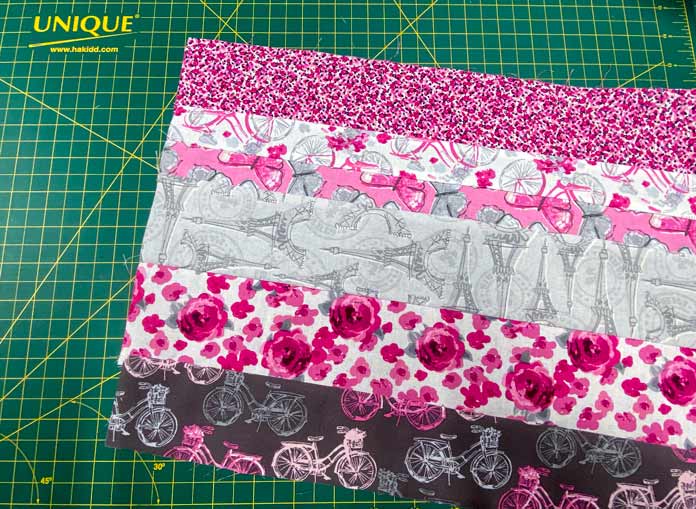 Six different fabric strips in different widths have been sewn together and sit on a green cutting mat. The fabrics are prints of pink, white, gray and maroon.