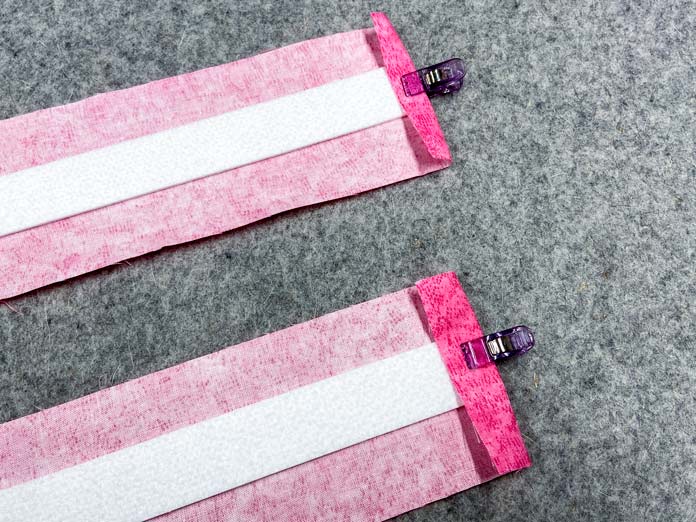 Two pink strips on fabric are shown laying right side down on a grey pressing mat. A narrow strip of white interfacing is clipped to the middle of each strip with a purple clip..