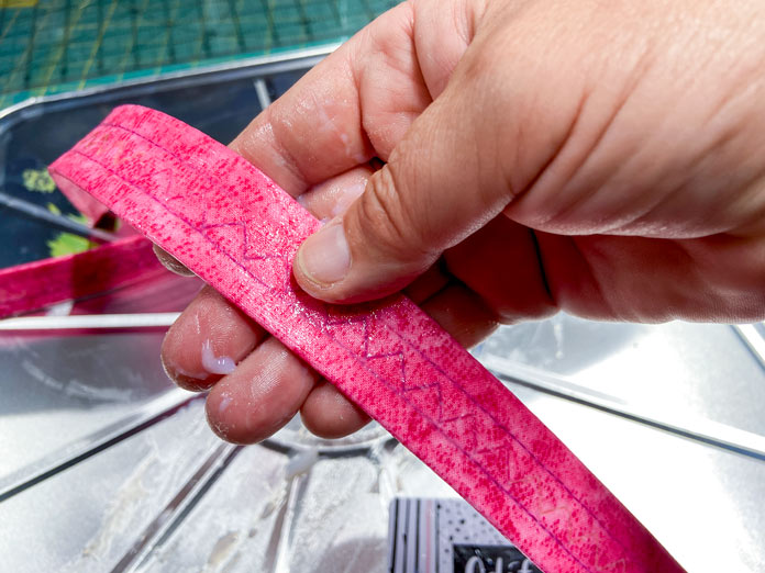 A hand holds a pink bag handle and rubs a gel coating into the fabric. A silver tray is in the background.