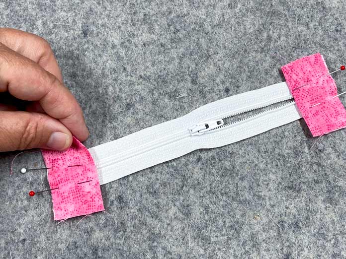 A hand holds a small pink square of fabric that has been pinned to one end of a white zipper. There is a second square at the other end of the zipper. The zipper sits on top of a grey pressing mat.