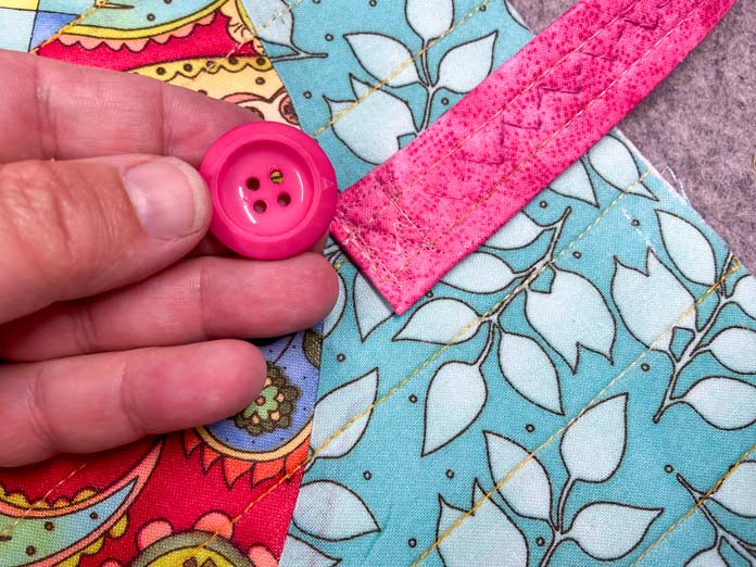 A hand holds a large pink button in front of a pink handle that has been sewn onto a multicolored quilted panel.