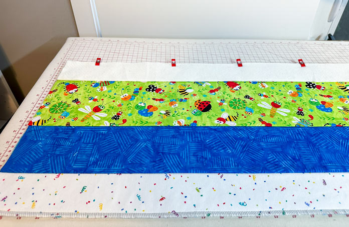 A quilt started with a white strip, blue strip and print (bottom to top). On the print raw edge is the fabric, batting, and backing all clipped together neatly aligning all the raw edges, with only the batting and clips showing.