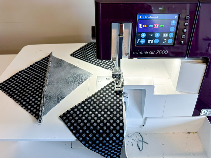 A white and burgundy serger with an extension table setup. Serging black and gray metallic triangular fabrics together bad sides together, so the bold decorative looper threads show.