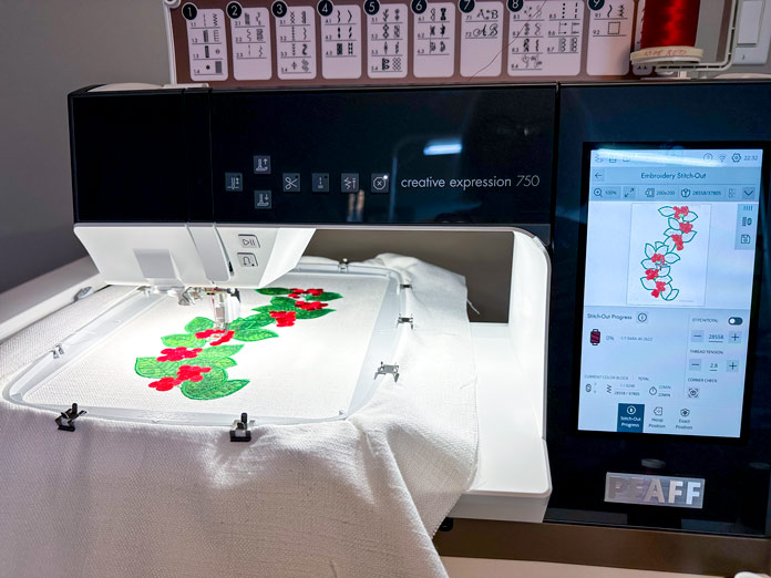 A black and white sewing and embroidery machine embroidering a green holly applique and red berry endless design in a 260X200 hoop on cream fabric.