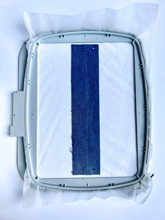 A hooped sample prepared for ribbon stitch embroidery using the PFAFF creative icon 2. A strip of denim is centered and pinned over stabilizer and white fabric inside the Elite Hoop 260 x 200 mm, ready for in-the-hoop ribbon stitching. This setup demonstrates the preparation stage before adjusting the hoop.