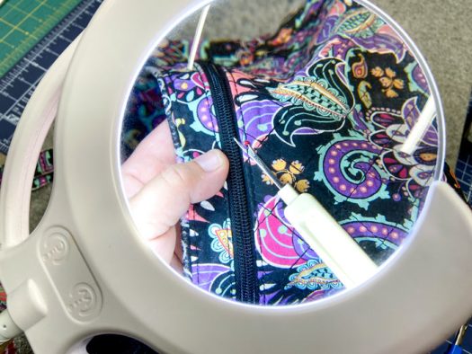 A hand holding a multicolored purse and a white seam ripper can be seen through a magnifying lamp lens.