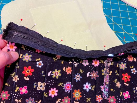 A hand holds a piece of black fabric with a zipper pinned to its top edge. A piece of light green fabric is seen in the background, lying on a green cutting mat.