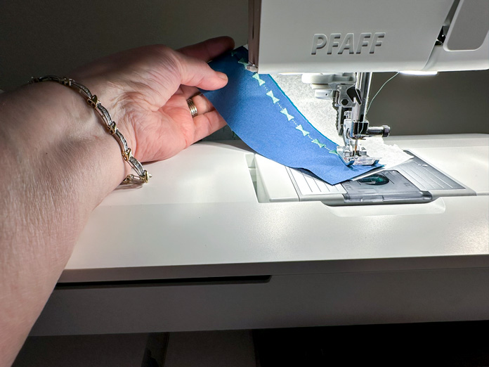 A blue piece of fabric, on top of a green (hidden) stitching a decorative seam along the edge of the fabric in green thread. White stabilizer showing out the side.