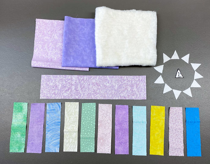 Cut out pieces of fabric in blues, greens, pinks and yellows and batting, with a small triangle template cut from freezer paper are laid out on a surface; Fairfield Quilter's 80/20 Quilt Batting