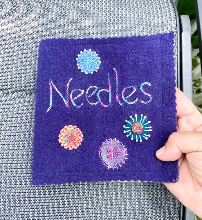 A hand holds a needle book made of purple wool with multicolored embroidery designs on the front with a gray seat cushion in the background; DMC Embroidery Floss, DMC Cotton Perle Thread