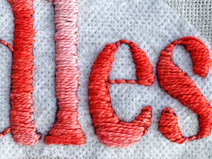 A close-up photo of a word embroidered with orange variegated DMC perle cotton and a satin stitch; DMC Charles Craft Monaco Needlework Fabric 28ct 20" x 24" - Tea Dyed, DMC Magic Paper, DMC Pearl Cotton Size 8 (80m) Thread Balls