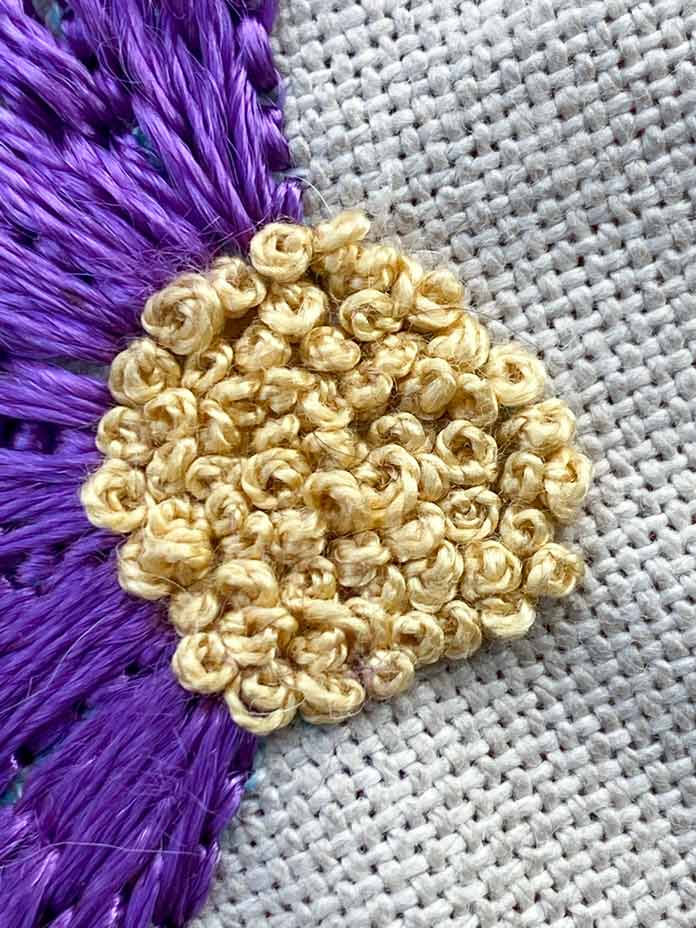 A close-up photo of the center of the purple flower shows how a gold-colored DMC perle cotton is used to stitch a bunch of small French knots close togther to fill in the area. The beige Charles Craft Monaco Needlework Fabric is in the background.