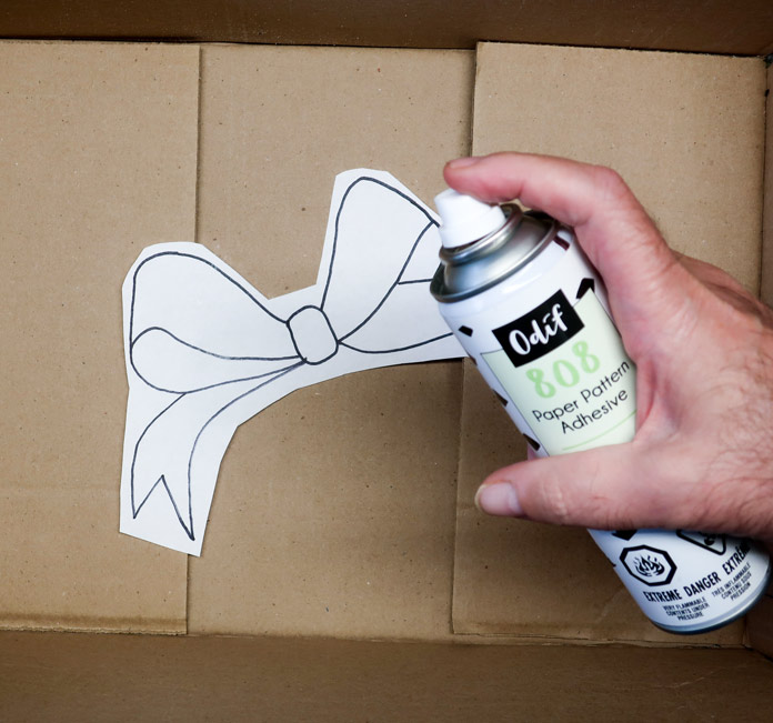 A bow paper template in a cardboard box, with a hand holding a can of spray adhesive positioned above; Odif 606 Spray and Fix No-sew Fusible Adhesive Web, Odif 808 Spray and Fix Temporary Adhesive for Paper Patterns, Odif 404 Spray and Fix Permanent Repositionable Adhesive for Craft Material, Komfort KUT Ruler Cutter, Gütermann 7-pc MCT Sew-all 100m Thread Set – Christmas