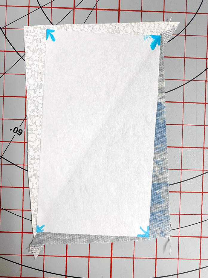 A rectangle piece of freezer paper is ironed to the right side of the Half Rectangle Triangle unit and the corner arrows are lined up with the seams on the unit. Gütermann Thread, SCHMETZ Quilting Needles, UNIQUE Sewing Fast Fade Fabric Marker, Omnigrid Ruler, OLFA Rotary Cutter, Komfort Kut Rotating Cutting Mat, Fabric Creations Cotton Fabric