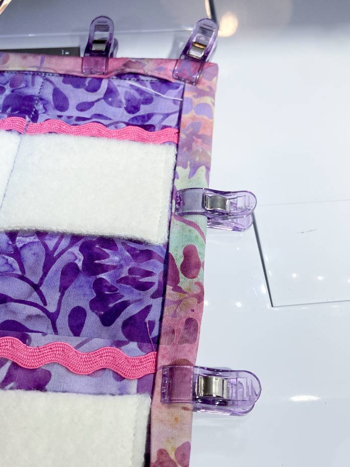 Four purple plastic clips hold pink batik binding in place on the edge of the purple batik fabric needle roll.