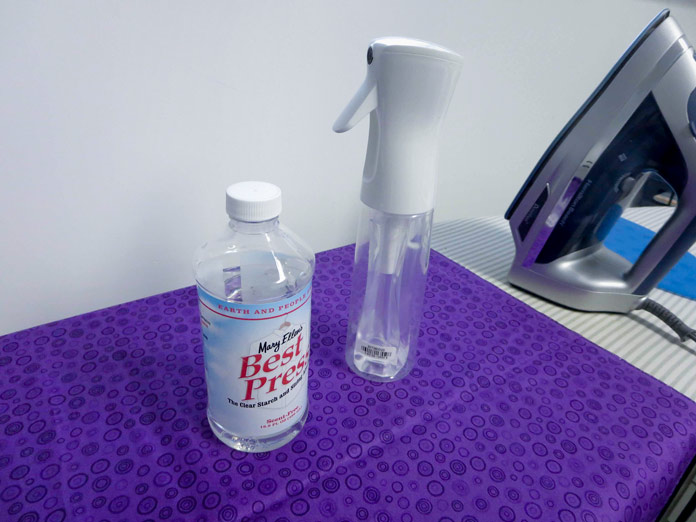Mary Ellen’s Best Press refill bottle and spray bottle sitting on wrinkled purple fabric next to an iron.