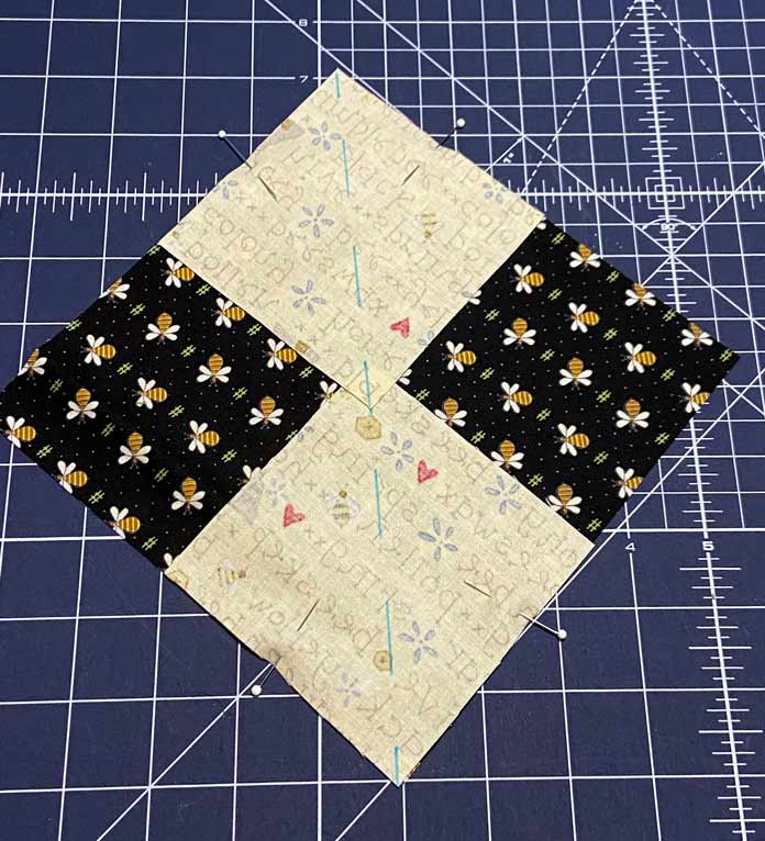 Two small squares of yellow fabric are pinned to opposite corners of a larger black square of fabric. The blue line drawn on the back of the yellow fabric squares is visible, and a blue cutting mat is in the background; OLFA Double Sided Cutting Mat (navy blue)